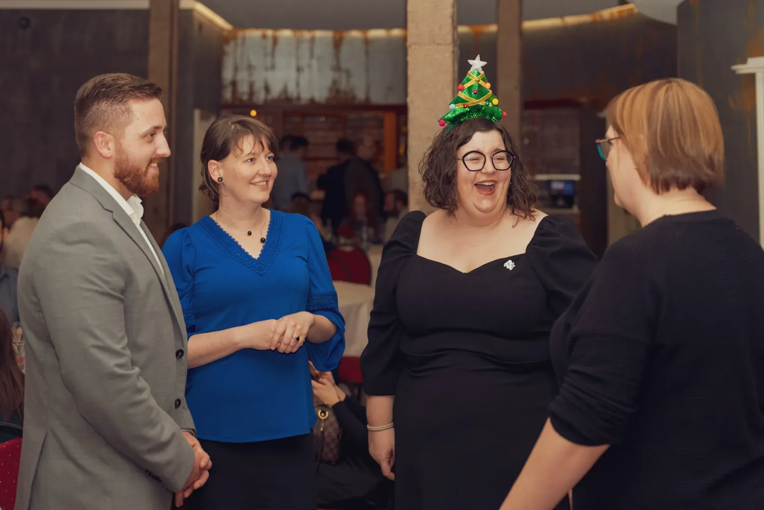 Eventfotograf Weihnachtsfeier: vierköpfige Gästerunde lacht gemeinsam, eine Teilnehmerin mit Weihnachtsbaum-Fascinator