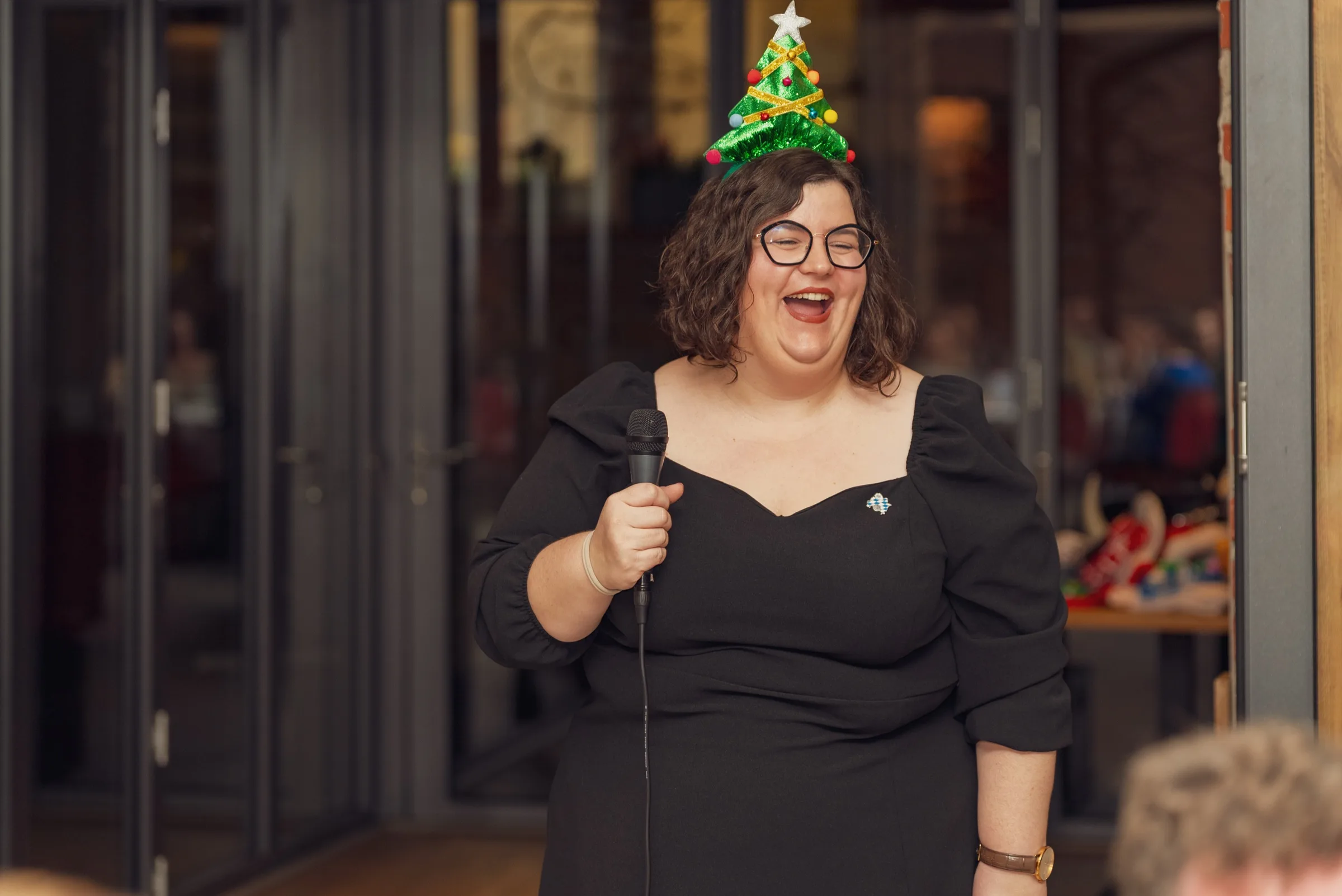 Eventfotograf Weihnachtsfeier: Moderatorin mit Weihnachtsbaum-Fascinator spricht ins Mikrofon
