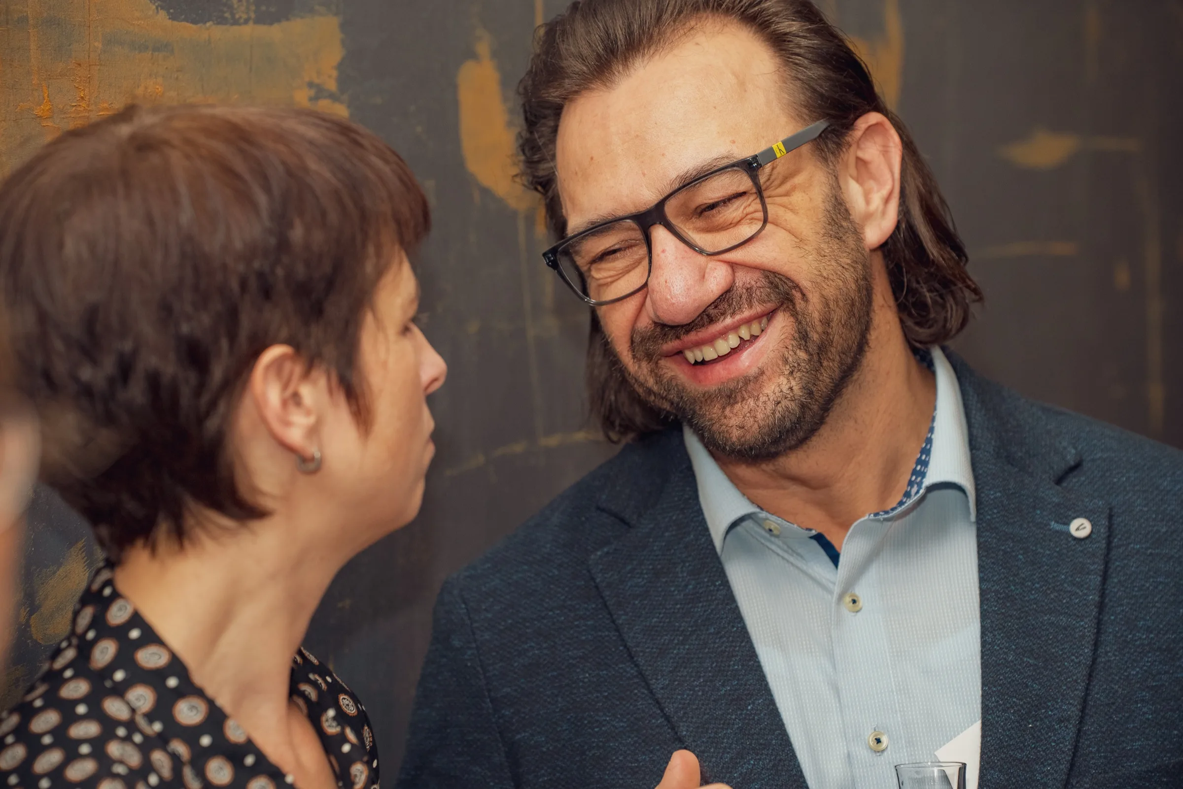Eventfotografie Firmenfeier: Gast lacht herzlich im Gespräch vor rustikaler Wand